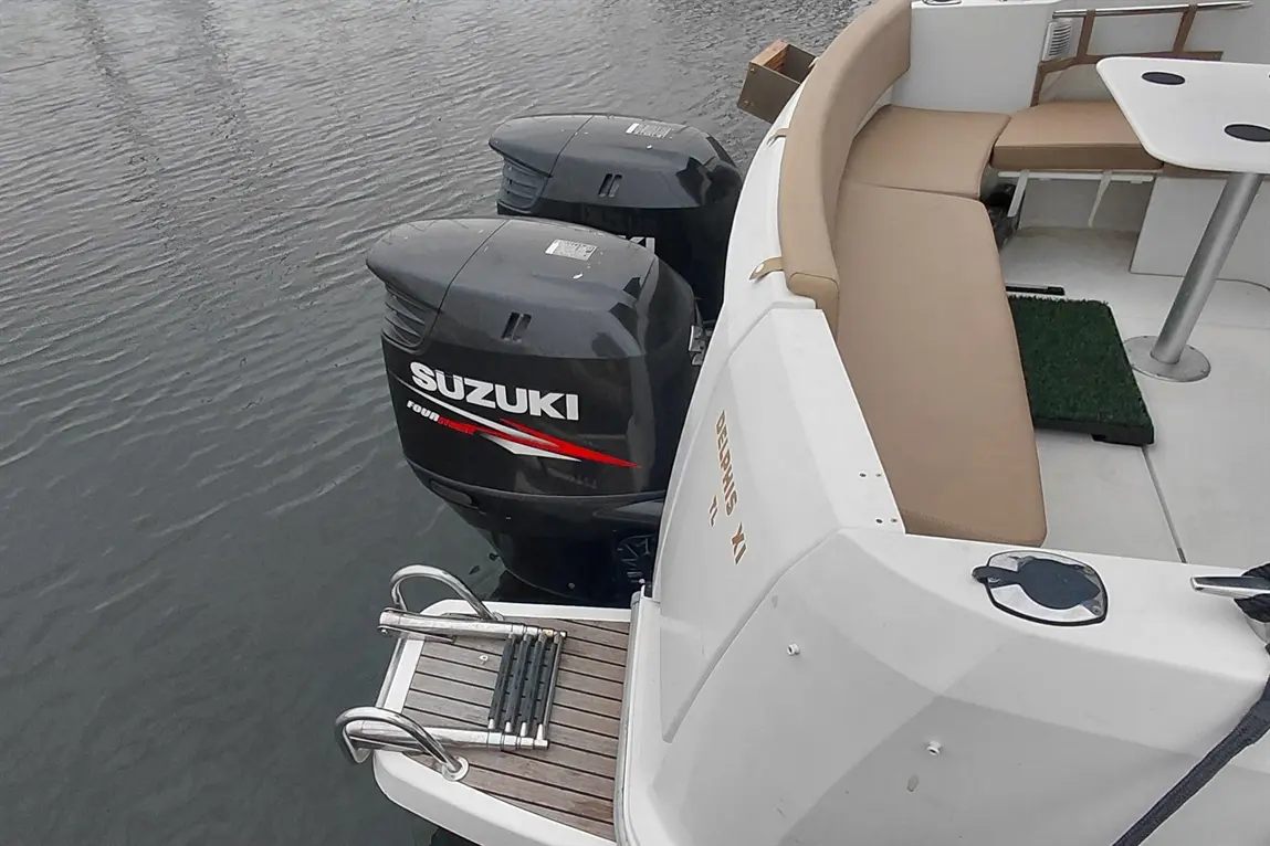 Twin Suzuki outboard motors are mounted on a boat, near beige seating and a table, with rippling water in the background.