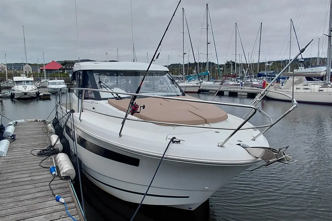 A white boat docked by a wooden pier, with a beige sunbed and fishing rod, surrounded by various boats in the marina.