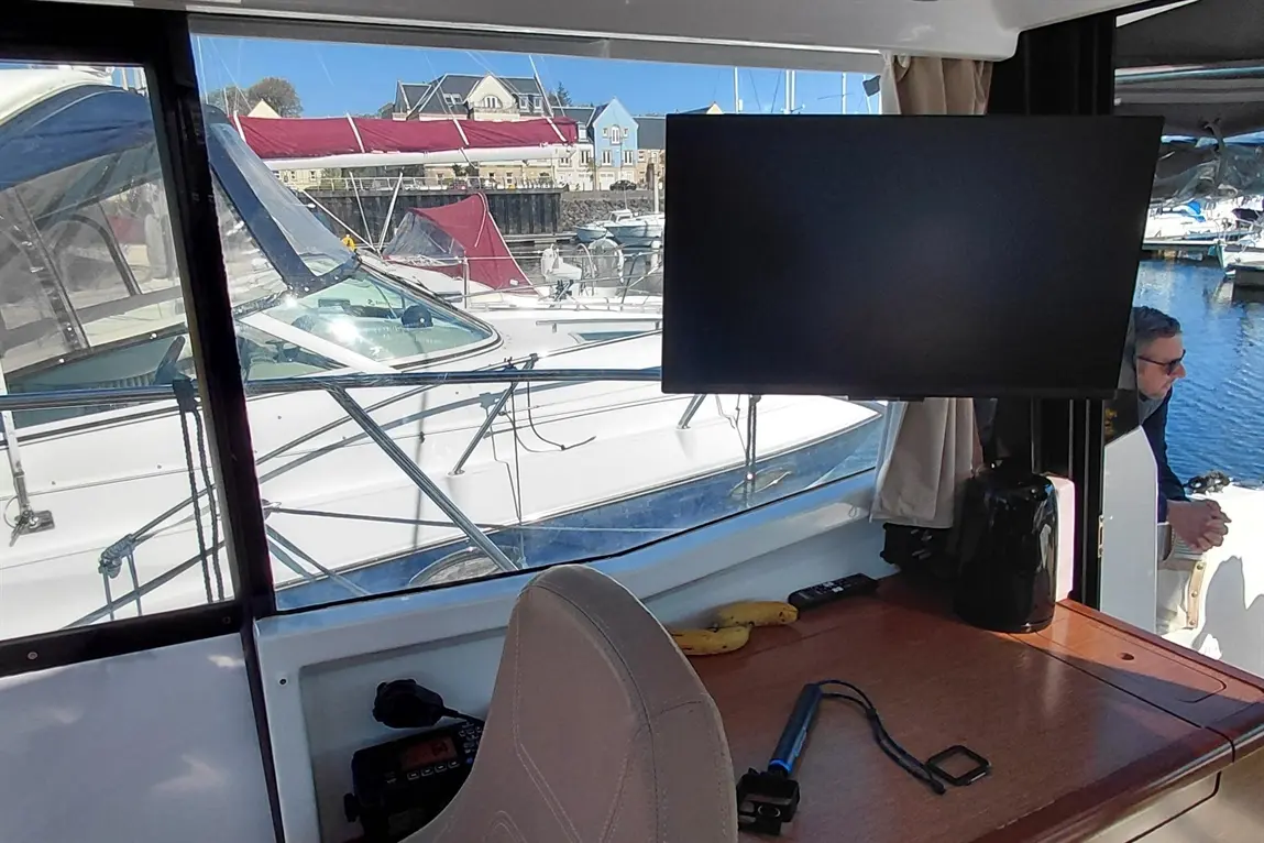 View from a boat interior showing a TV screen, boat controls, and bananas on a table, with a marina in the background.