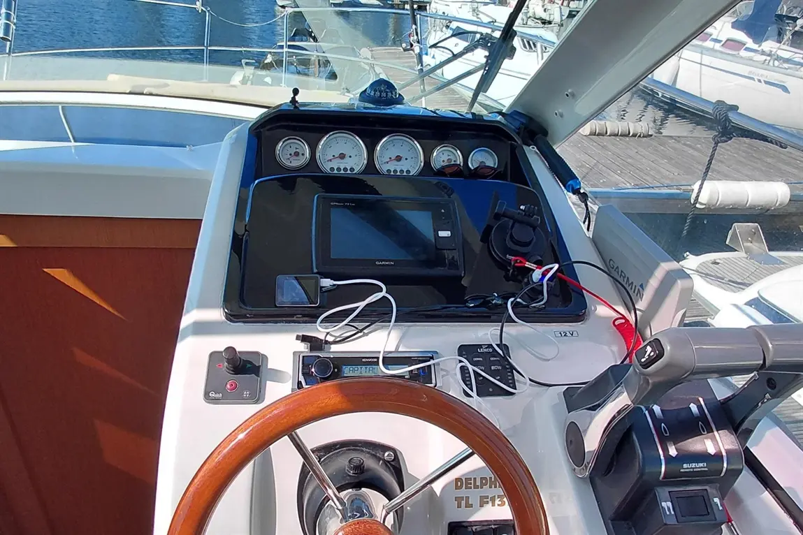 Boat dashboard with a wooden steering wheel, Garmin display, gauges, and controls, overlooking a marina. Text: "DELPHIN TL F13".