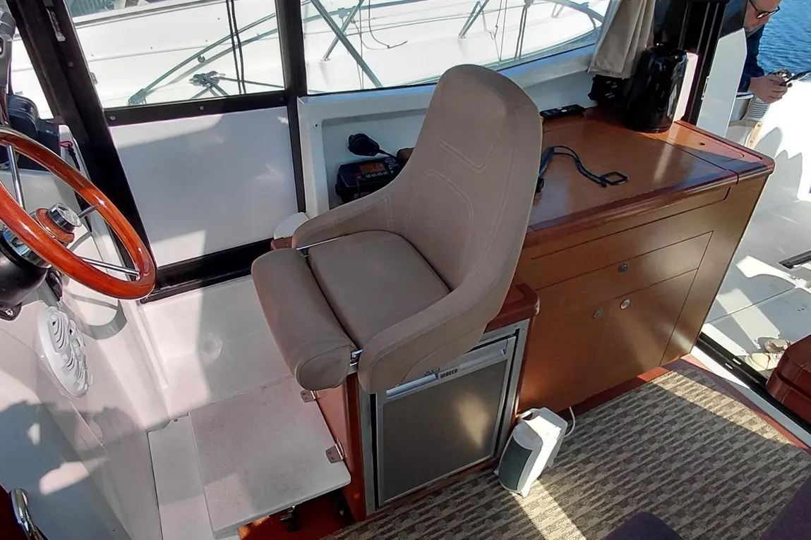 A comfortable helm chair beside a wooden console, steering wheel, and controls, with a view of the water outside.