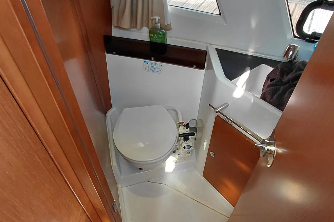 Compact boat toilet with a white toilet seat, a sink, and a towel on the side; natural light enters through a window.