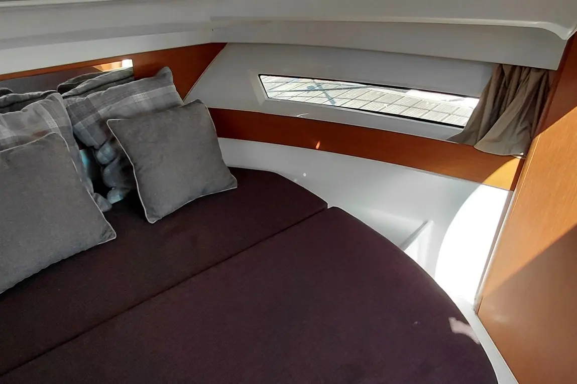 Cosy interior of a yacht cabin featuring a double bed with grey cushions and a window showing the dock outside.