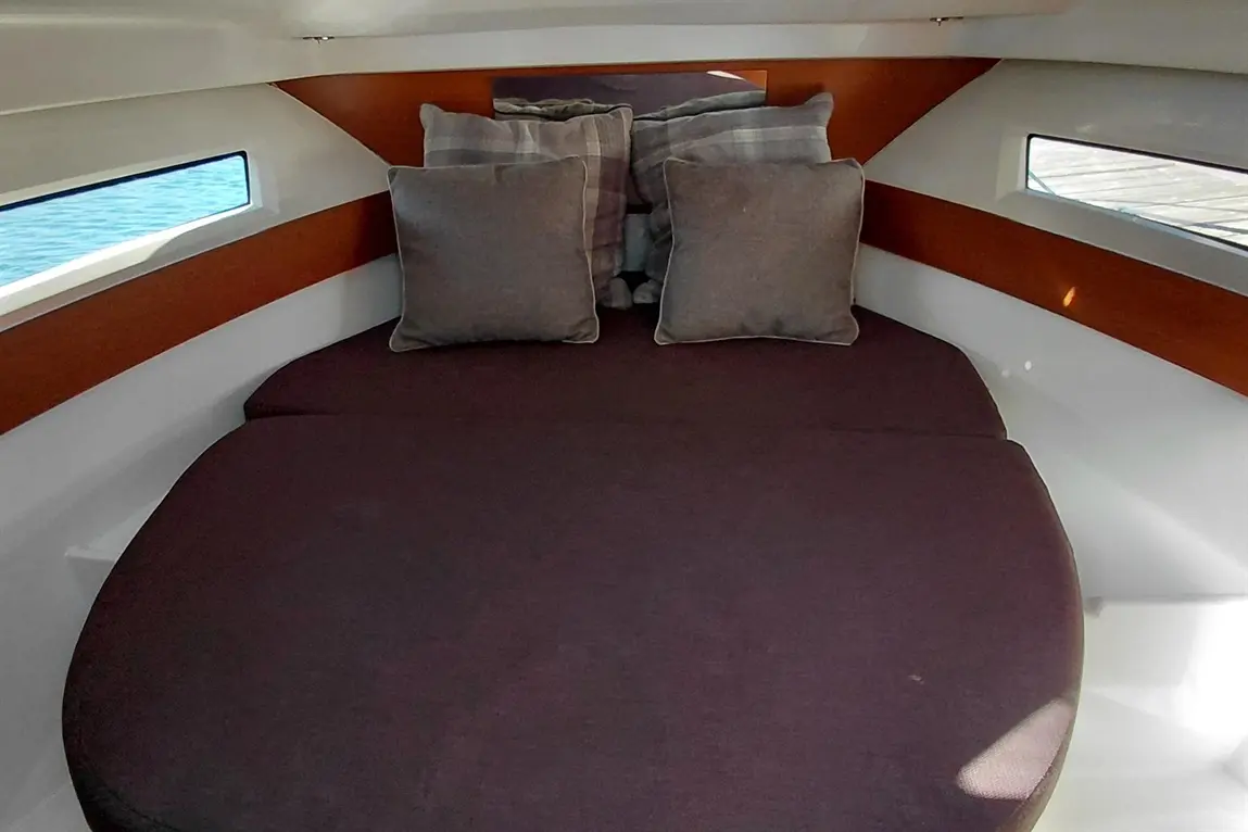 A cosy cabin interior featuring a rounded bed with dark cushions and windows showing a view of the water outside.