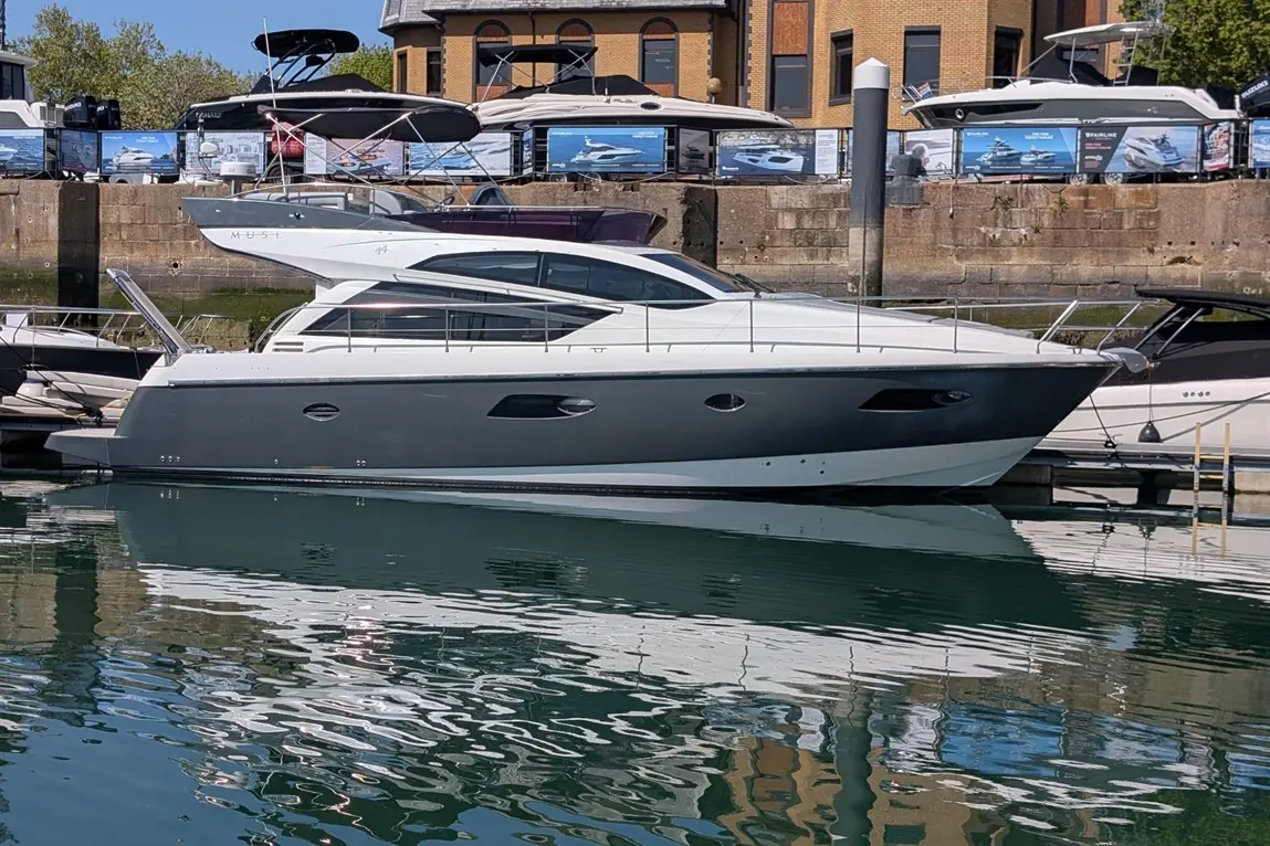 A modern grey and white motor yacht docked in calm water, reflecting buildings and other boats in the marina.