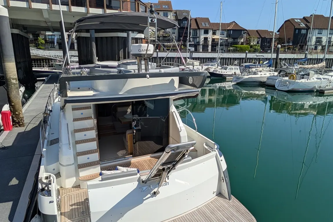 A luxury boat in a marina, featuring steps to the cabin and reflections in clear water, with various moored boats in background.