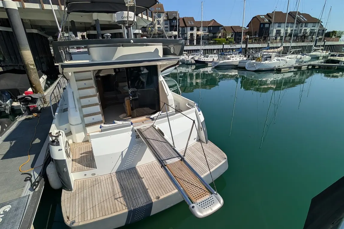 A motorboat docked in a marina, featuring a large rear deck, steps leading inside, and reflection on calm water.