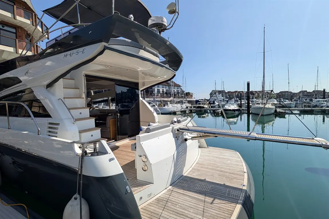 A luxury yacht named "MUSE" docked at a marina, with boats and a clear blue sky reflected in the calm water.