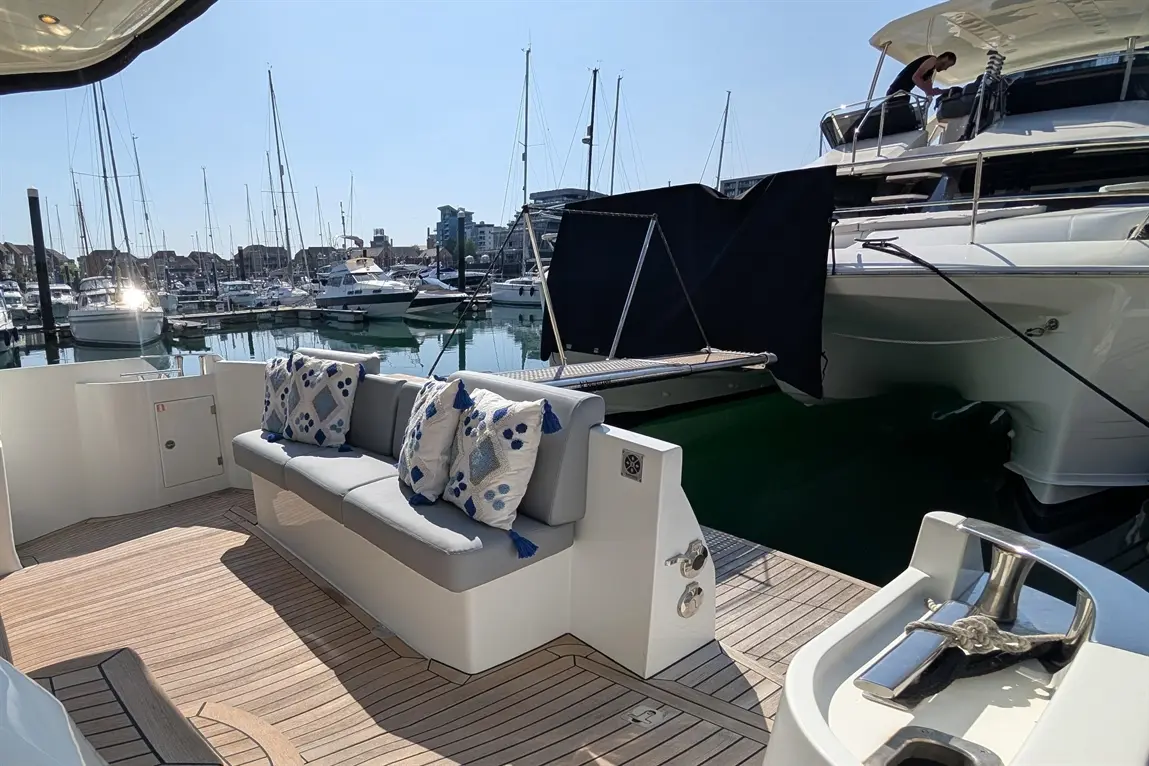 Luxury yacht deck with grey seating, decorative cushions, and a marina view featuring moored boats and a clear blue sky.
