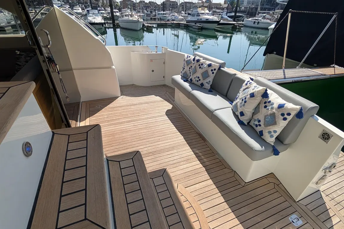 A sunlit yacht deck features a grey cushioned seating area with decorative pillows, overlooking a marina of boats.