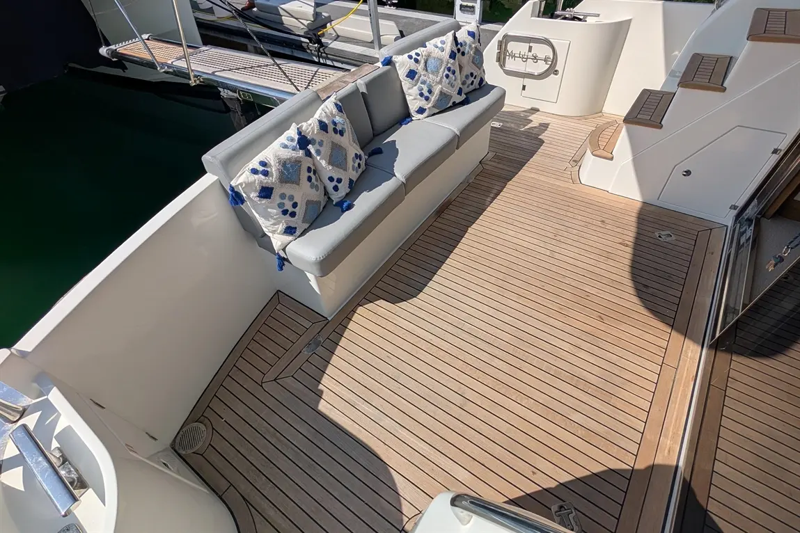 Luxury yacht deck featuring a grey cushioned bench with decorative pillows and wooden flooring, with a glimpse of water.