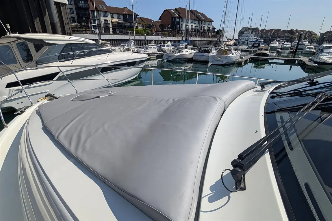 View from the bow of a boat showing a grey cover, adjacent boats, and reflections on a calm marina water under blue sky.