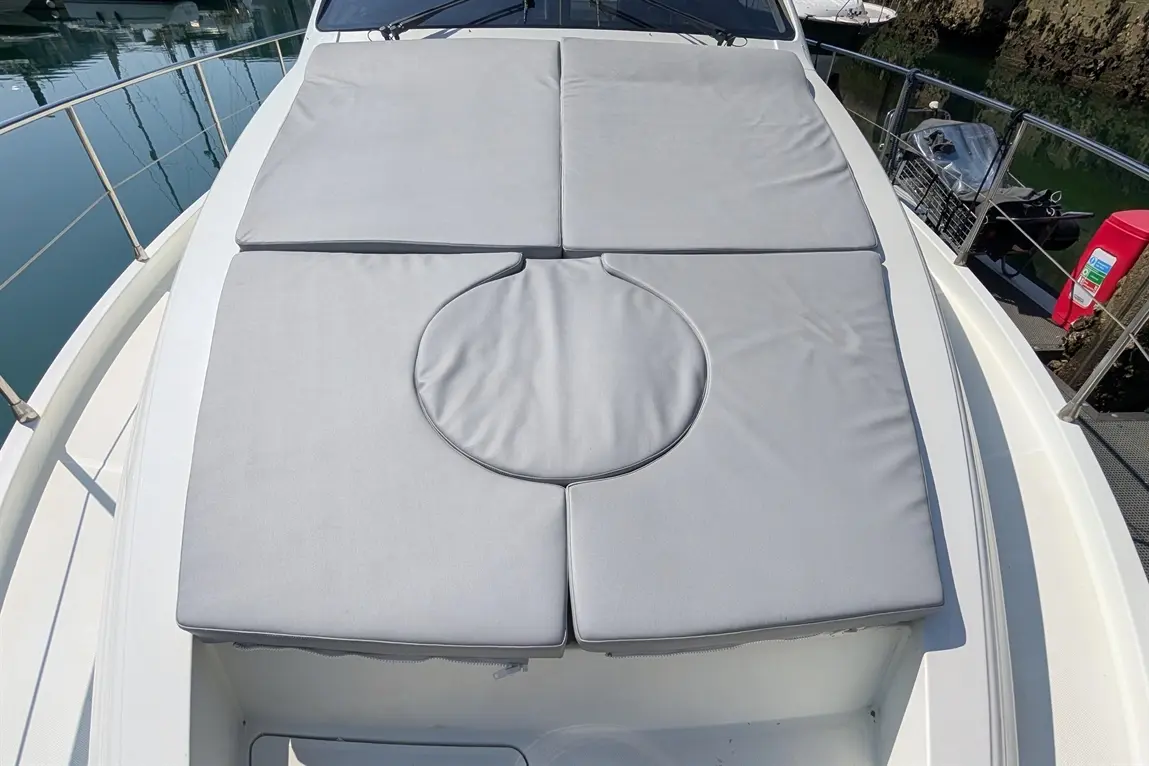 A grey padded sunbed on a boat, adjacent to calm waters, with a circular cutout for a sunshade or umbrella.