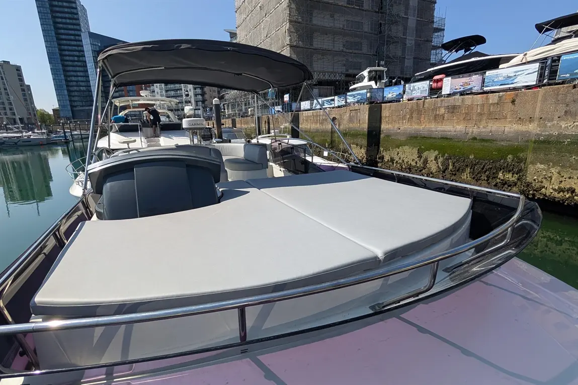 A sleek boat moored at a marina, featuring a large sunbed area and a view of high-rise buildings and docks in the background.