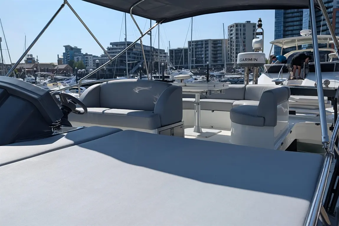 A modern boat cockpit features grey seating and table, with a view of a marina and city buildings in the background.