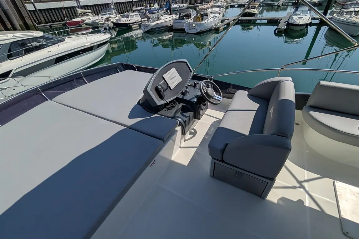 The helm of a modern boat with a steering wheel, cushioned seating, and a view of moored yachts in a marina.