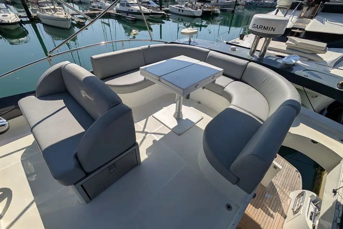 Stylish grey seating area on a boat, featuring a table, with boats docked in the marina reflecting on the water.