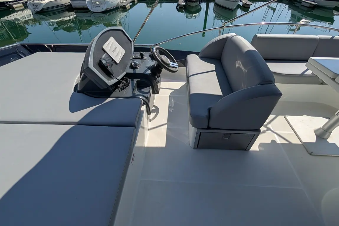 Interior of a boat showing a steering wheel, control panel, a cushioned seat, and reflections of boats in the water.