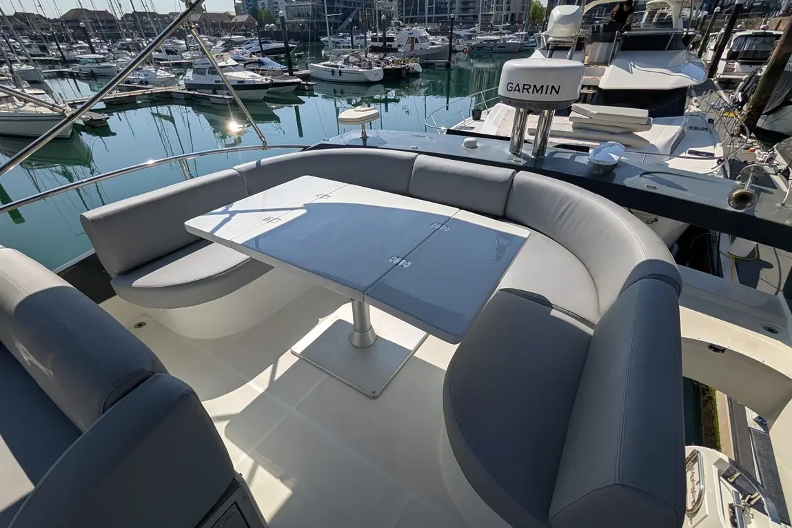 A modern boating deck features grey upholstered seating, a foldable table, and a Garmin device, overlooking a marina.