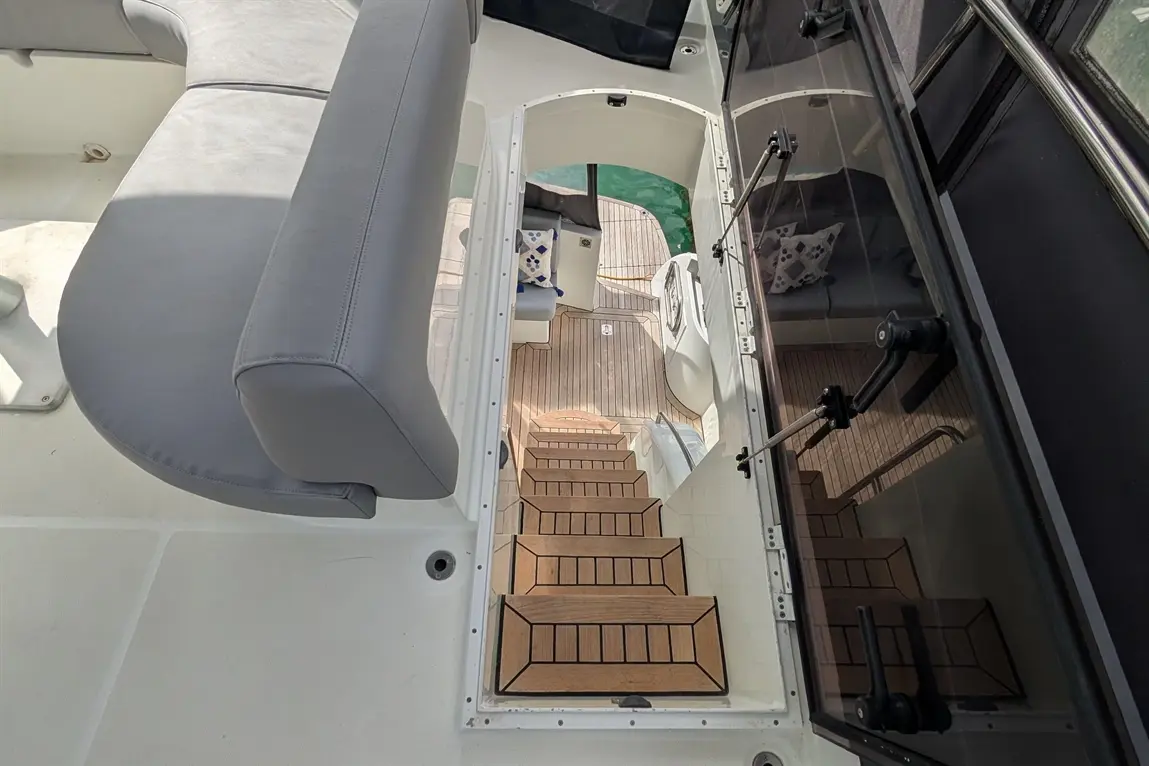 View of a yacht interior showing stairs leading down from the upper deck to the lower level, with grey seating and wood flooring.
