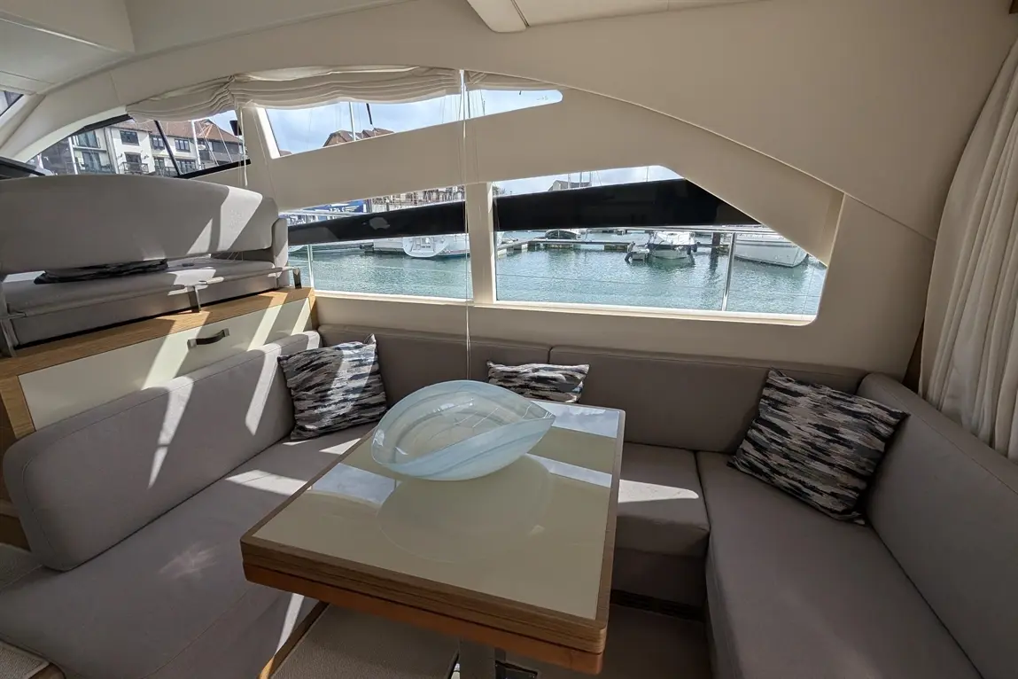 Interior of a yacht featuring a grey seating area, a glass table with a decorative bowl, and a marina view through large windows.