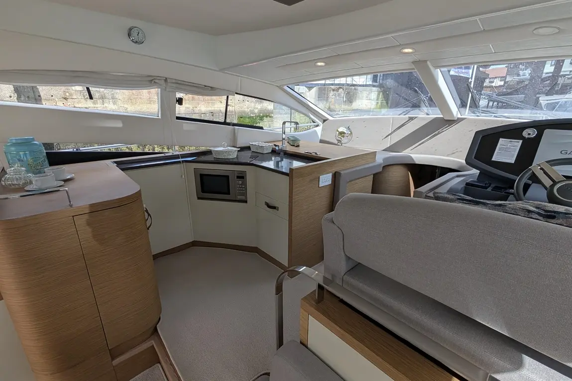 Modern boat interior with a kitchen area, microwave, and grey seating; large windows provide natural light and a scenic view.