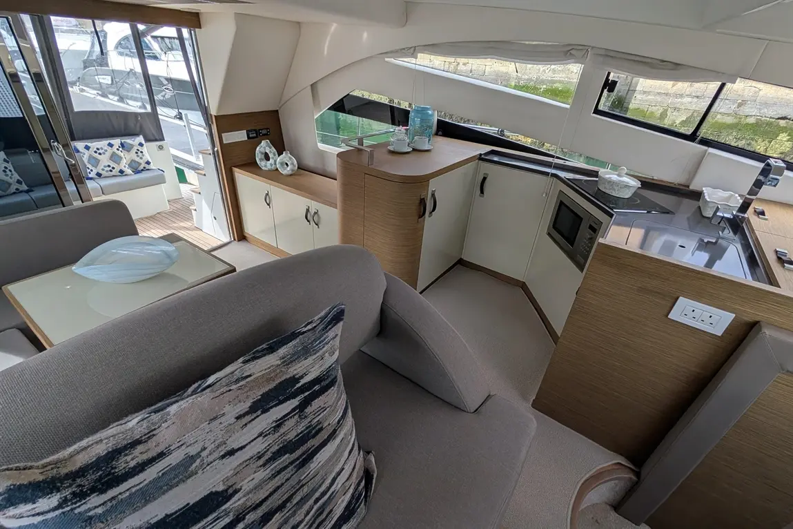 Interior view of a modern boat's lounge and kitchenette, featuring a sofa, table, and kitchen appliances.