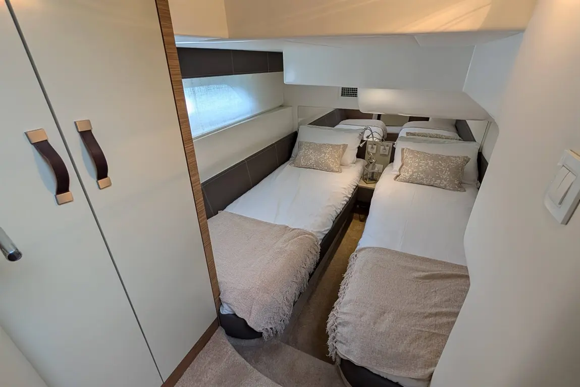 Compact bedroom with two single beds, light bedding, soft pillows, and a bedside table, featuring a window and cupboard.