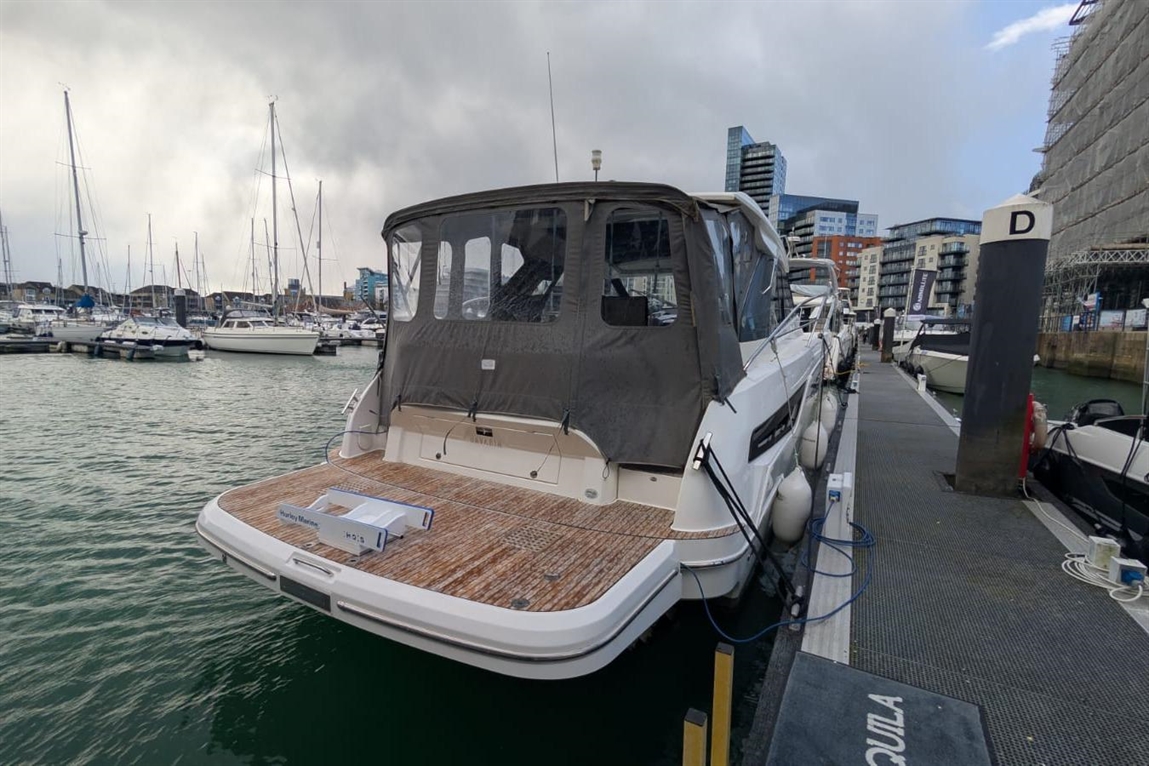 A spacious motorboat docked at a marina, surrounded by sailboats, with buildings and cloudy skies in the background.