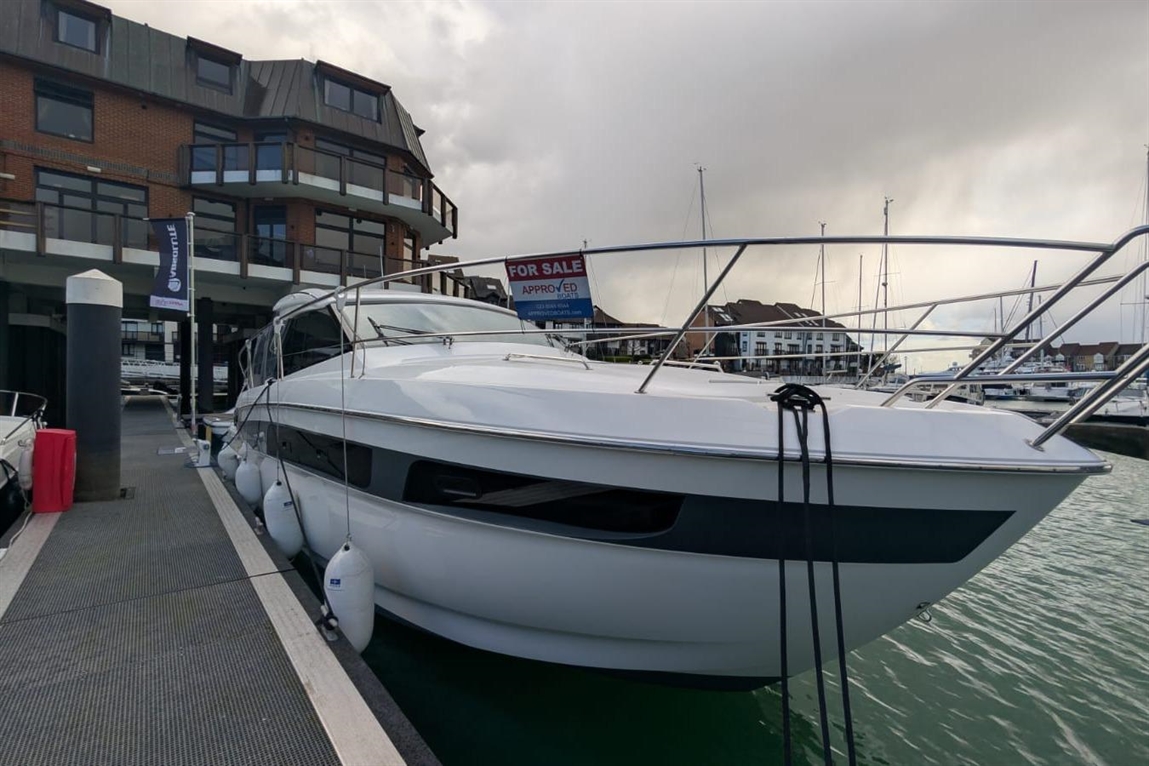 A modern white yacht docked at a marina, featuring a "FOR SALE" sign with the word "APPROVED" visible in the background.