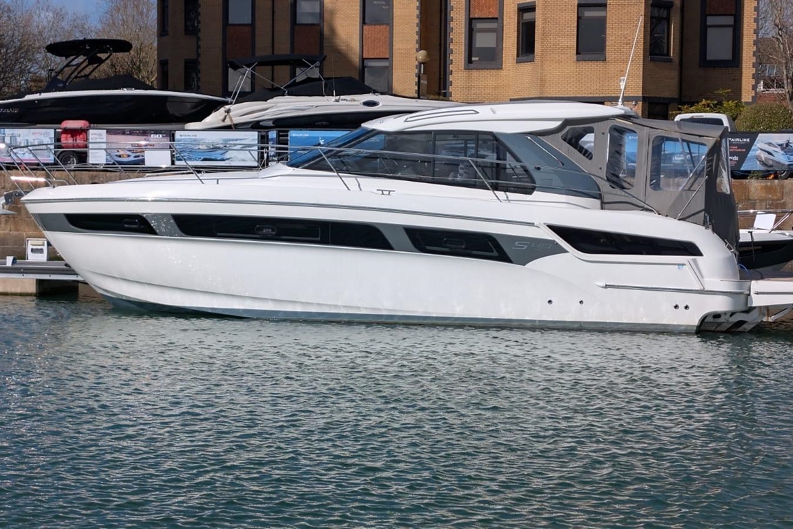 White luxury motorboat docked in water, showcasing sleek design and glass windows, with boats and buildings in the background.