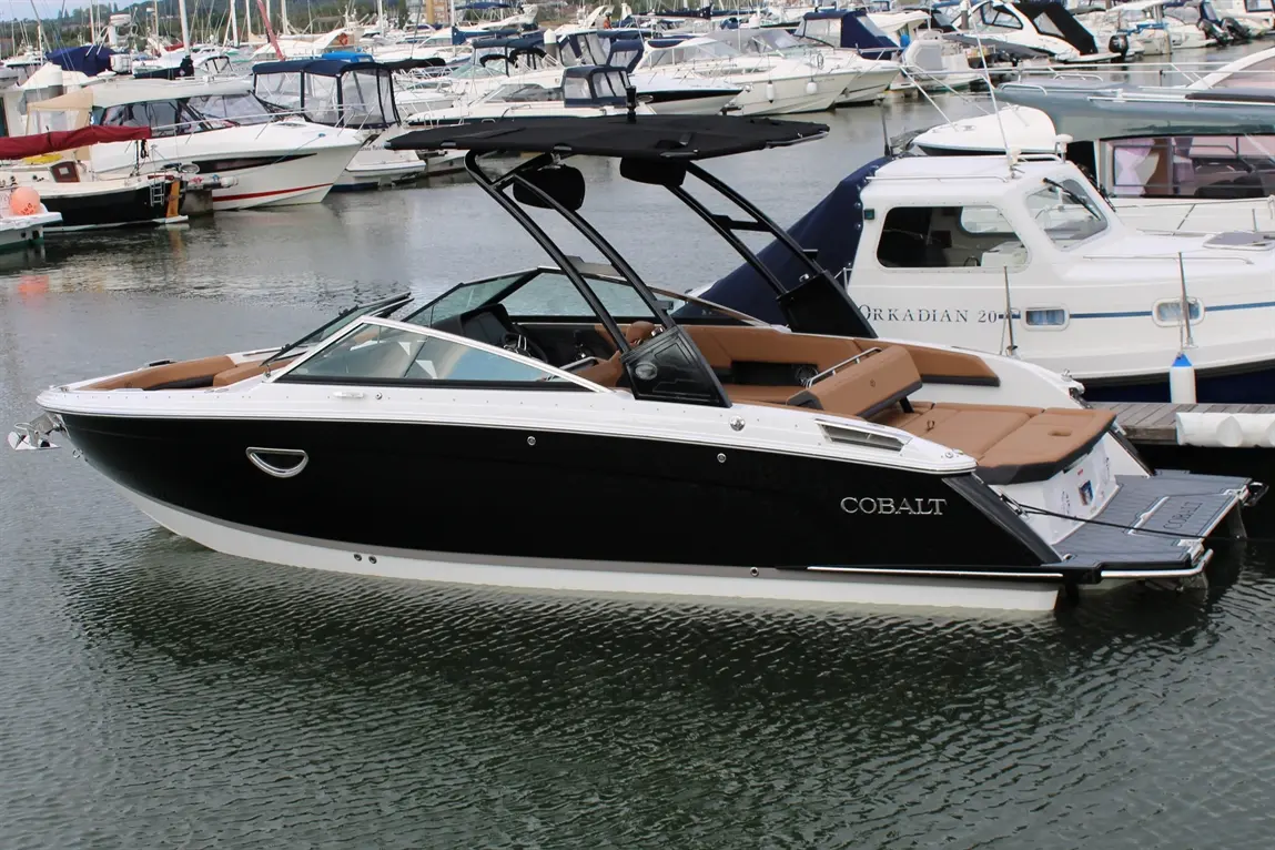 A sleek black Cobalt boat with tan seating moored in a marina, surrounded by various other boats and reflections on water.