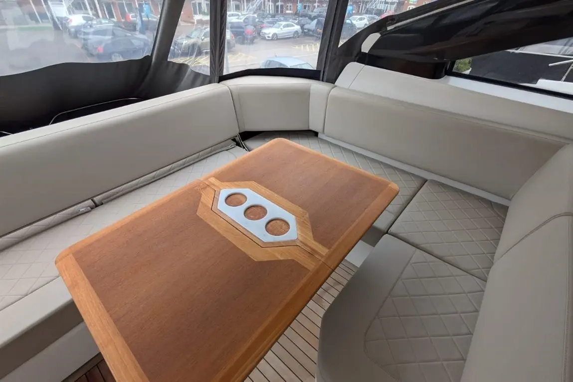 Interior of a modern boat featuring a wooden table and plush seating, with a view of parked cars outside through large windows.