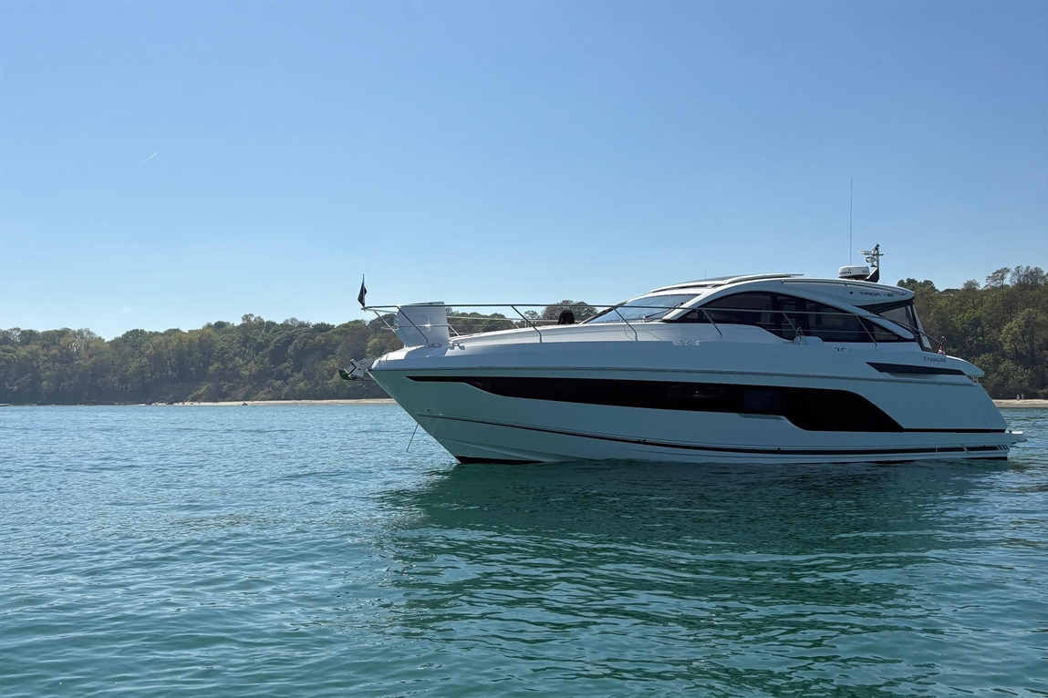 White luxury yacht anchored in clear water, with trees and a sandy beach in the background under a clear blue sky.