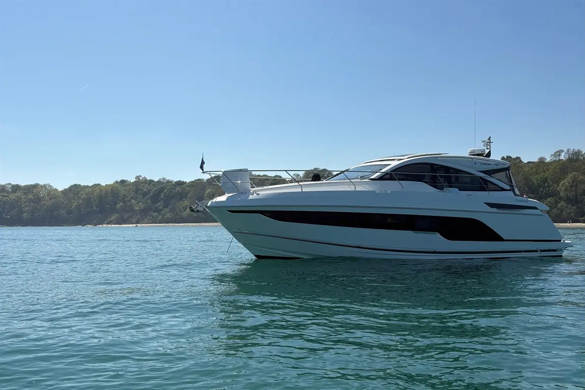 White luxury yacht anchored in clear water, with trees and a sandy beach in the background under a clear blue sky.