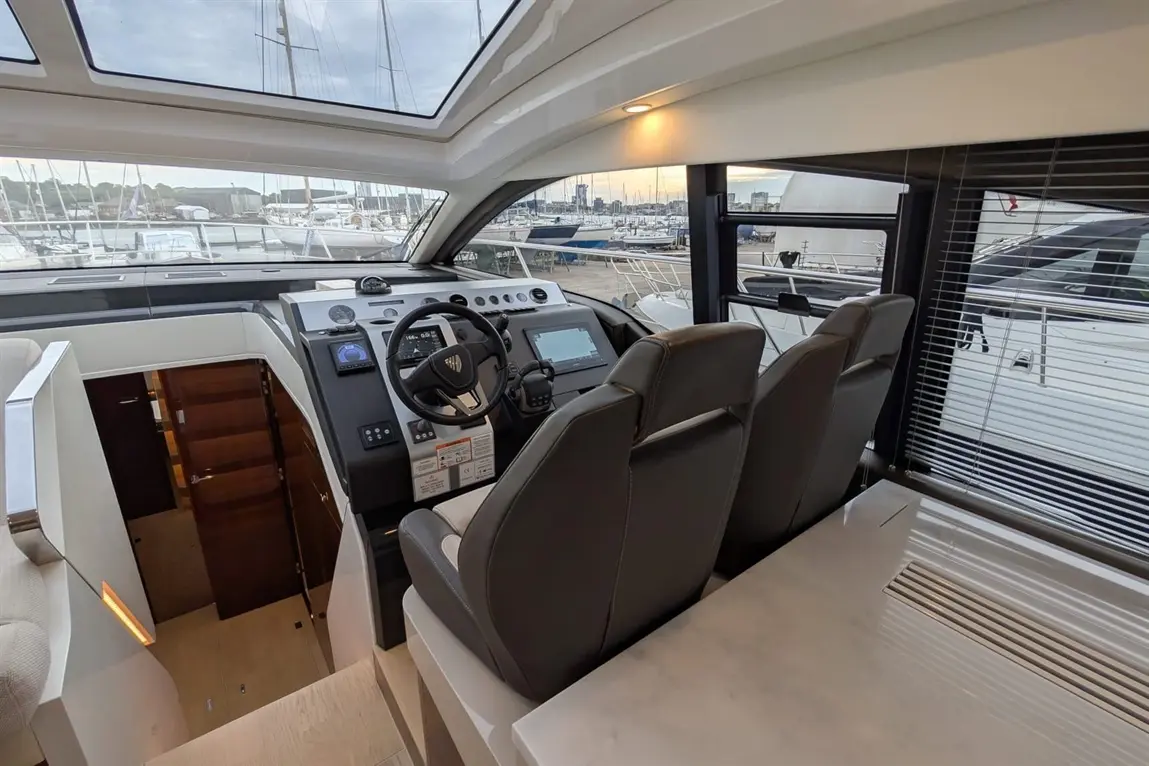 Modern yacht interior featuring a helm with controls, two grey captain's chairs, and a view of a marina through large windows.