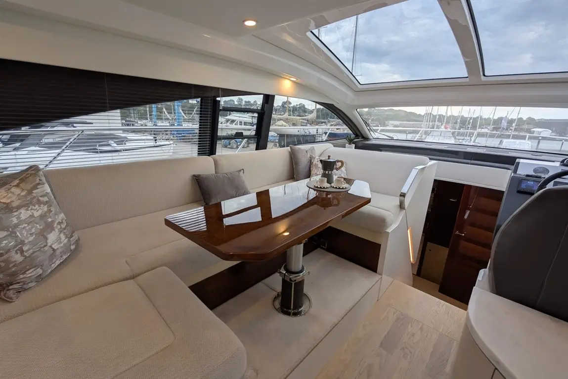 Interior of a yacht featuring a cream sofa, a polished wooden table, and scenic views of the marina through large windows.