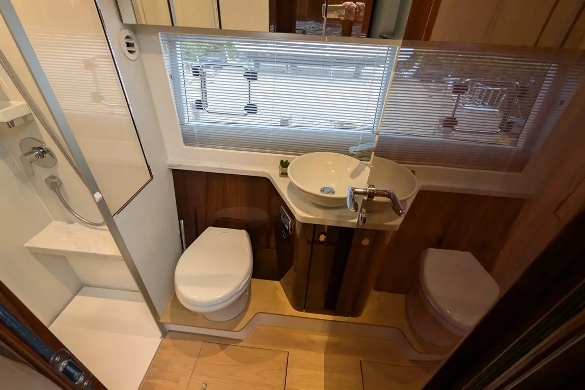 Compact bathroom with shower, modern sink, and toilet, featuring wooden accents and a window with blinds.