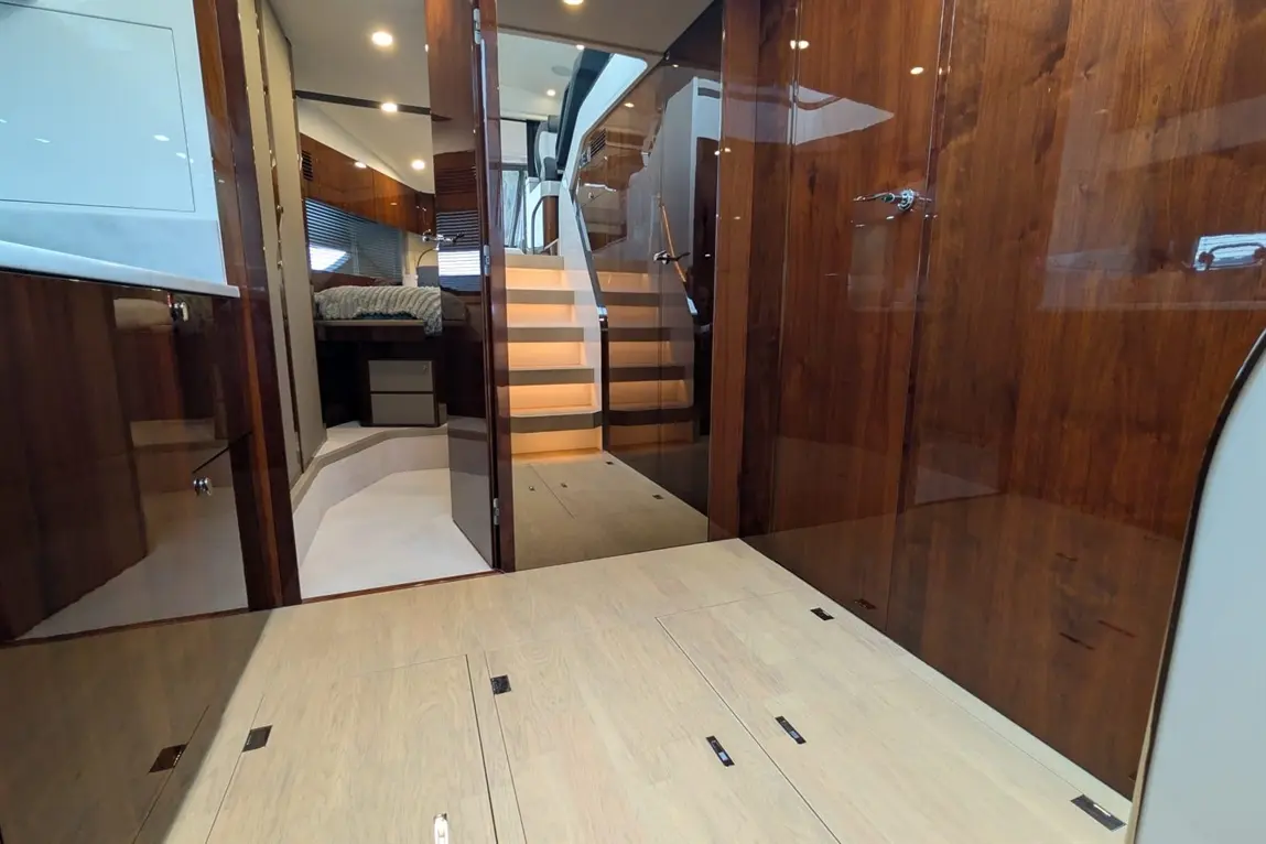 Modern yacht interior featuring polished wooden surfaces, stairs leading up, and a glimpse of a bedroom area with a quilted bed.