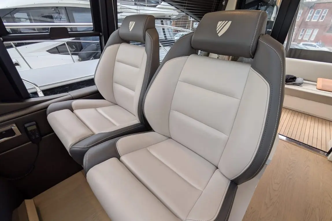 Two modern boat seats in grey and cream leather, with visible stitching, overlooking yachts through large windows.