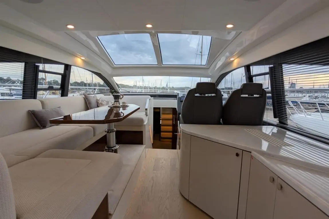 Interior of a modern yacht, featuring a dining area, two captain's seats, and panoramic windows overlooking a marina.