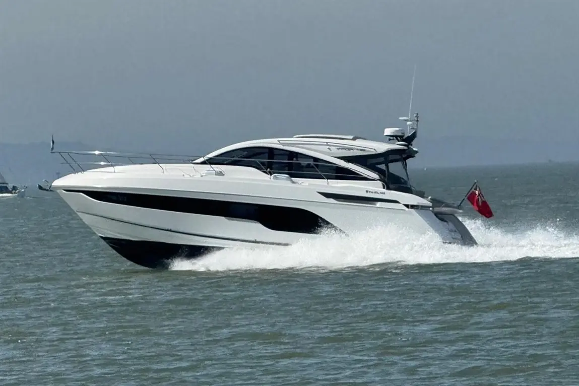 A sleek white motorboat speeds through water, creating waves, with a small red flag visible on the stern.
