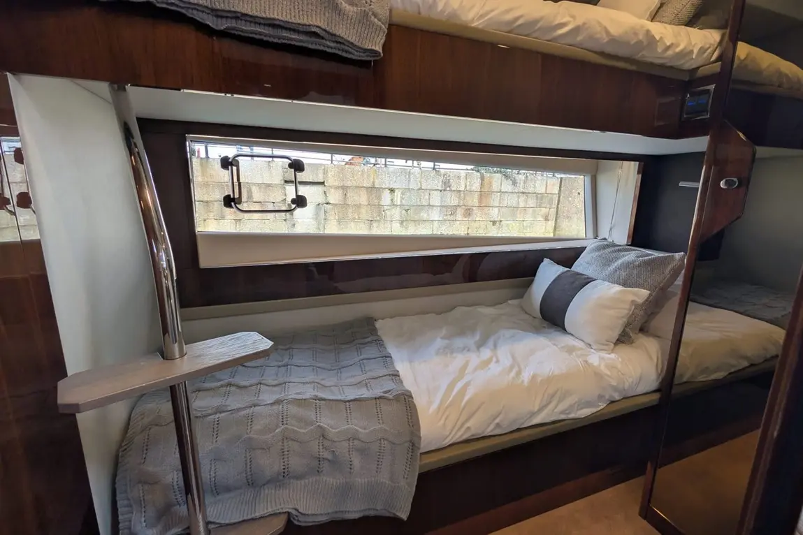 Interior of a boat cabin featuring a bed with grey throw, white pillows, and a window revealing a stone wall outside.
