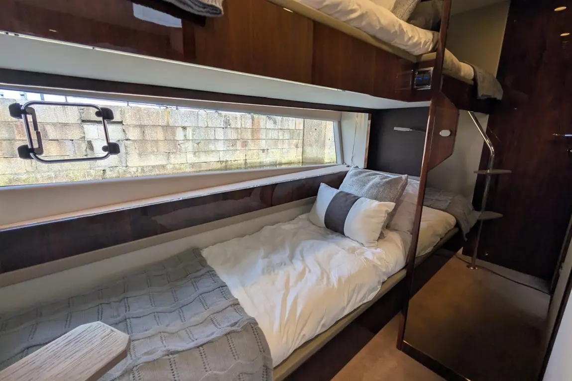 Interior view of a cosy cabin with a single bed, decorative pillows, and a ladder to a top bunk, featuring a window with a stone wall outside.