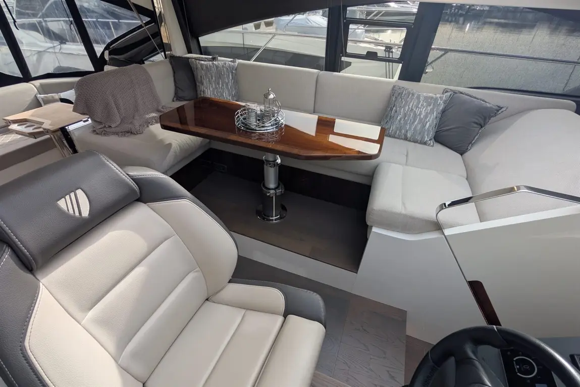 Luxurious boat interior featuring a seating area, polished wooden table, cushions, and a modern helm chair, with a marina view.
