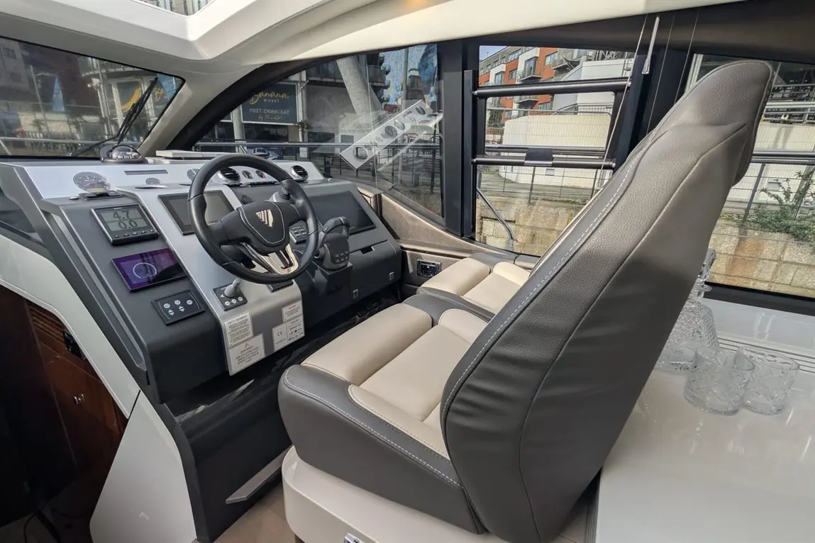 A modern boat cockpit featuring a steering wheel, digital screens, and a comfortable captain's chair, with a view of the marina.