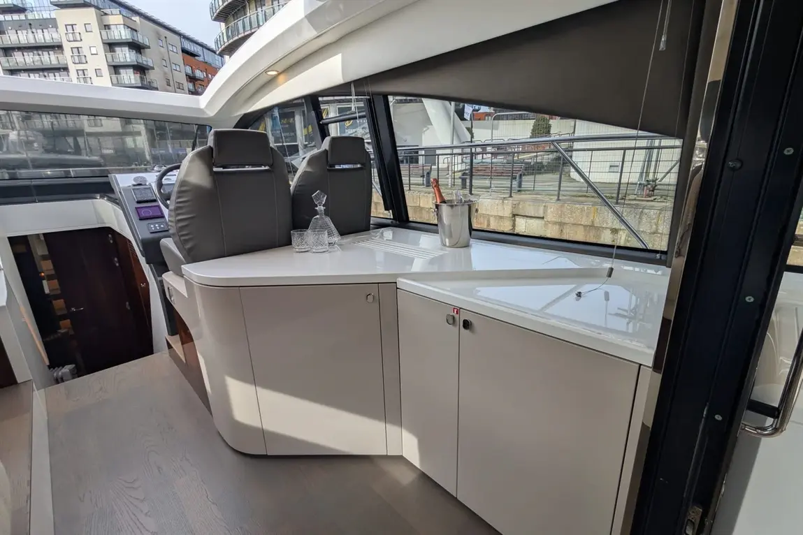 Interior of a luxury boat featuring grey captain's chairs, a bar area with glasses and a champagne bucket, and sunlight through large windows.