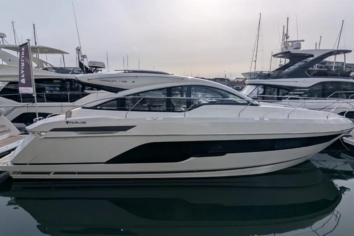 A sleek Fairline boat moored at a marina, reflecting on the water, with other yachts in the background on a cloudy day.