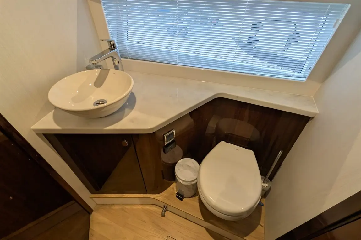Modern bathroom interior featuring a round sink, stylish tap, toilet, bin, and wooden flooring with sunlight through blinds.