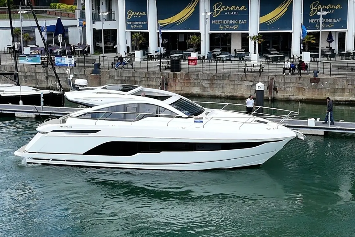 A sleek white Fairline yacht docked at Banana Wharf, with people seated nearby and staff preparing for service.